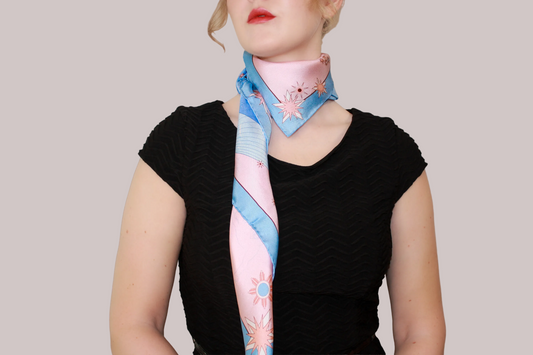A light blue and pink celestial patterned silk scarf, Gemini Lovers by The Line Atelier, tied around a model's neck over a black top, set against a solid taupe background.