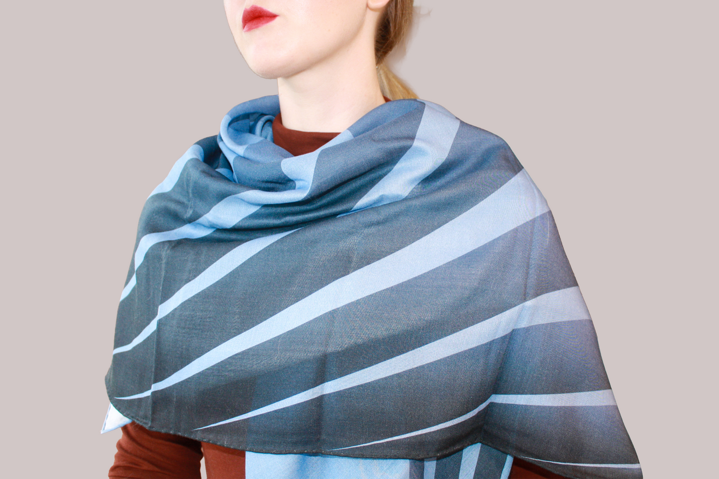 A woman wearing a blue and light blue striped shawl, Ice Valley silk sand wool by The Line Atelier, draped over her shoulders, styled with a rust-brown mock-neck top, a black belt, and dark gray trousers, set against a solid beige background.