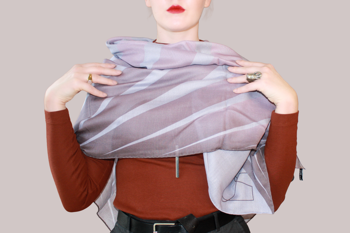 A woman wearing a blue and light blue striped shawl, Love Potion nr 9 silk sand wool by The Line Atelier, draped over her shoulders, styled with a rust-brown mock-neck top, a black belt, and dark gray trousers, set against a solid beige background.