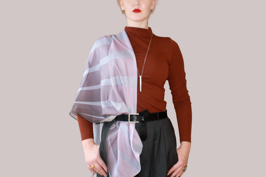 A woman wearing a blue and light blue striped shawl, Love Potion nr 9 silk sand wool by The Line Atelier, draped over her shoulders, styled with a rust-brown mock-neck top, a black belt, and dark gray trousers, set against a solid beige background.