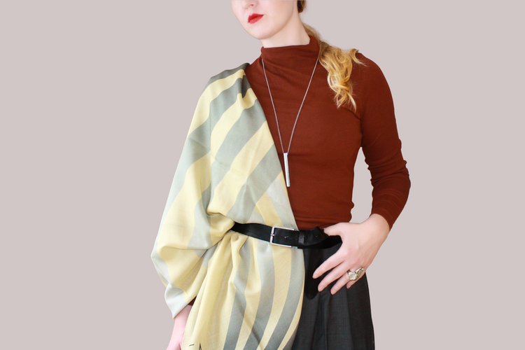 A woman wearing a blue and light blue striped shawl, Willow Grove silk sand wool by The Line Atelier, draped over her shoulders, styled with a rust-brown mock-neck top, a black belt, and dark gray trousers, set against a solid beige background.