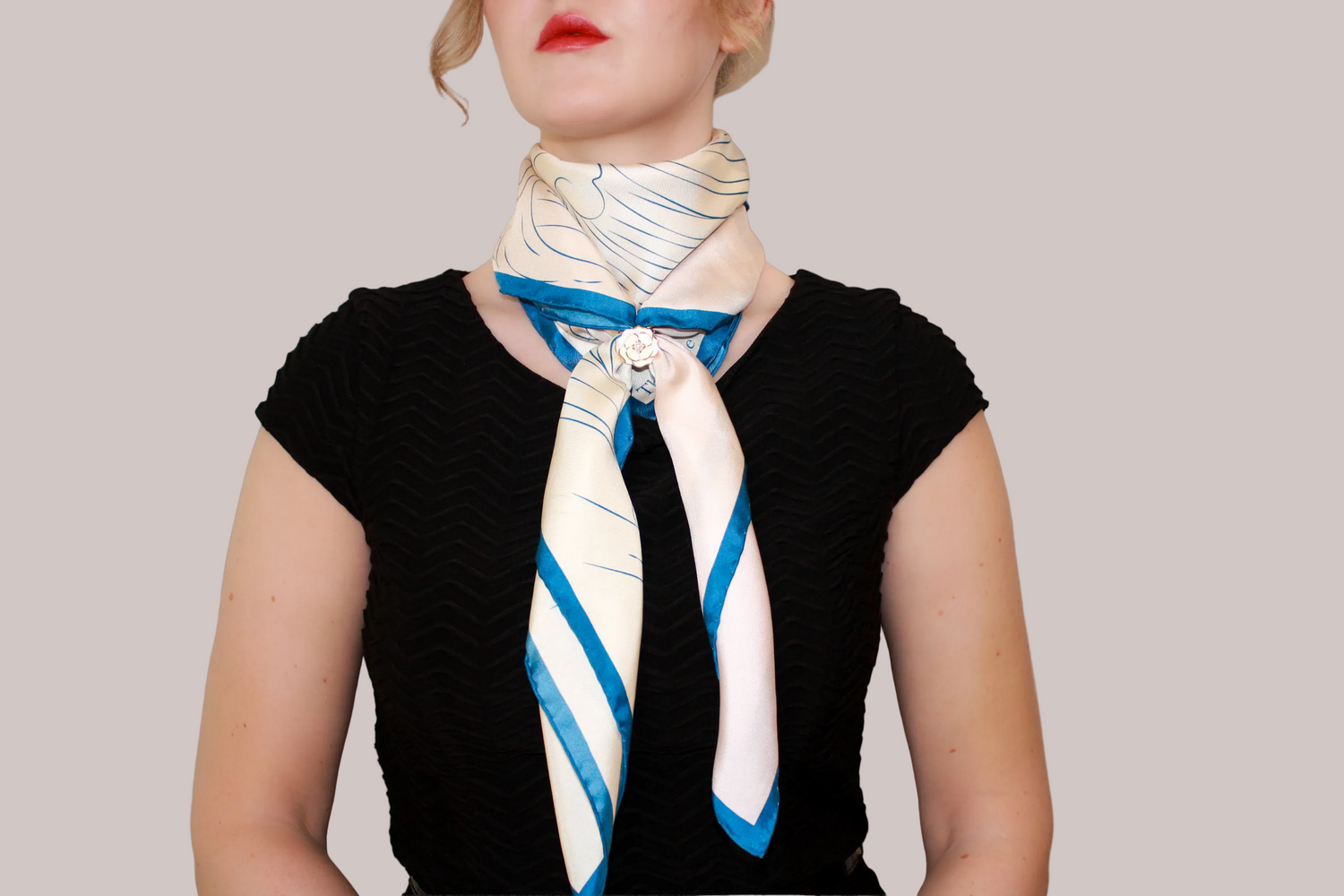 A close-up of a model wearing a cream-colored silk scarf Halcyon Seas with a blue and brown feather pattern, styled over her shoulders.