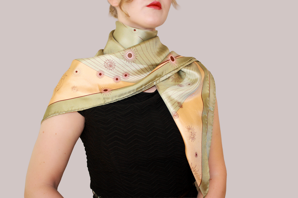 A woman wearing a silk scarf, Courage of Leo by The Line Atelier, tied around her shoulders, featuring a light green and peach Leo constellation pattern with small red floral accents, set against a solid taupe background.