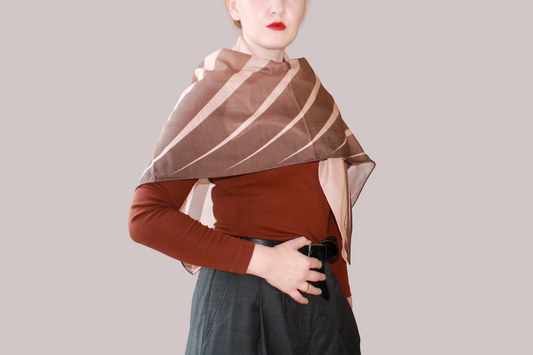 A woman wearing a blue and light blue striped shawl, Bronze Sandrift silk sand wool by The Line Atelier, draped over her shoulders, styled with a rust-brown mock-neck top, a black belt, and dark gray trousers, set against a solid beige background.