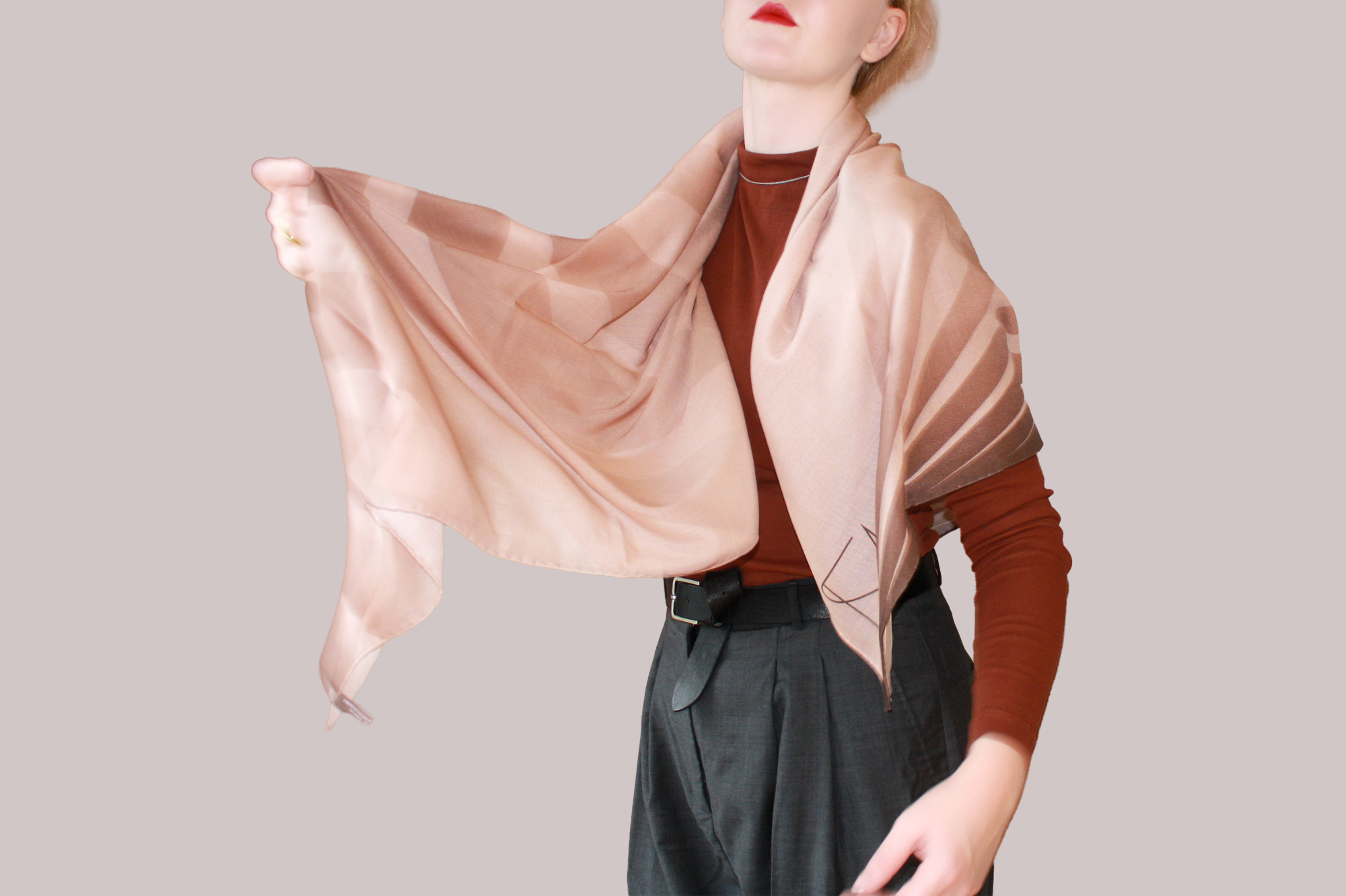 A woman wearing a blue and light blue striped shawl, Bronze Sandrift silk sand wool by The Line Atelier, draped over her shoulders, styled with a rust-brown mock-neck top, a black belt, and dark gray trousers, set against a solid beige background.