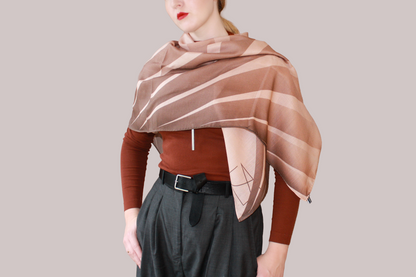 A woman wearing a blue and light blue striped shawl, Bronze Sandrift silk sand wool by The Line Atelier, draped over her shoulders, styled with a rust-brown mock-neck top, a black belt, and dark gray trousers, set against a solid beige background.