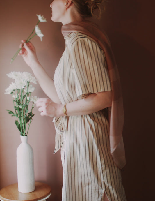 Model wearing Bronze Sandrift Wool & Silk Scarf holding a flower with a vase of flowers on a table against a brown background