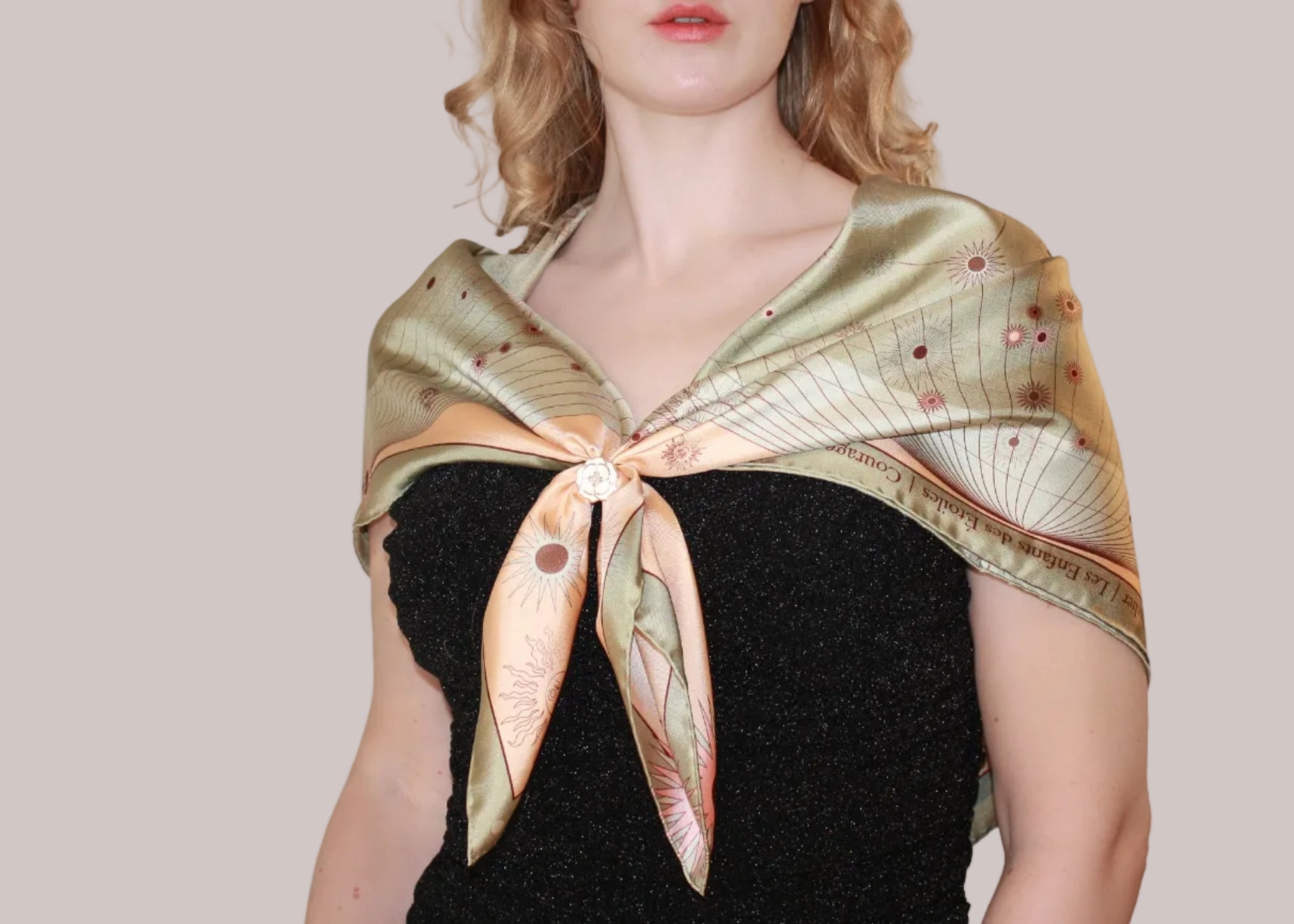 A woman wearing a gold-toned silk scarf with a celestial Leo pattern and small pink accents, tied around her shoulders over a black textured top against a solid light beige background.