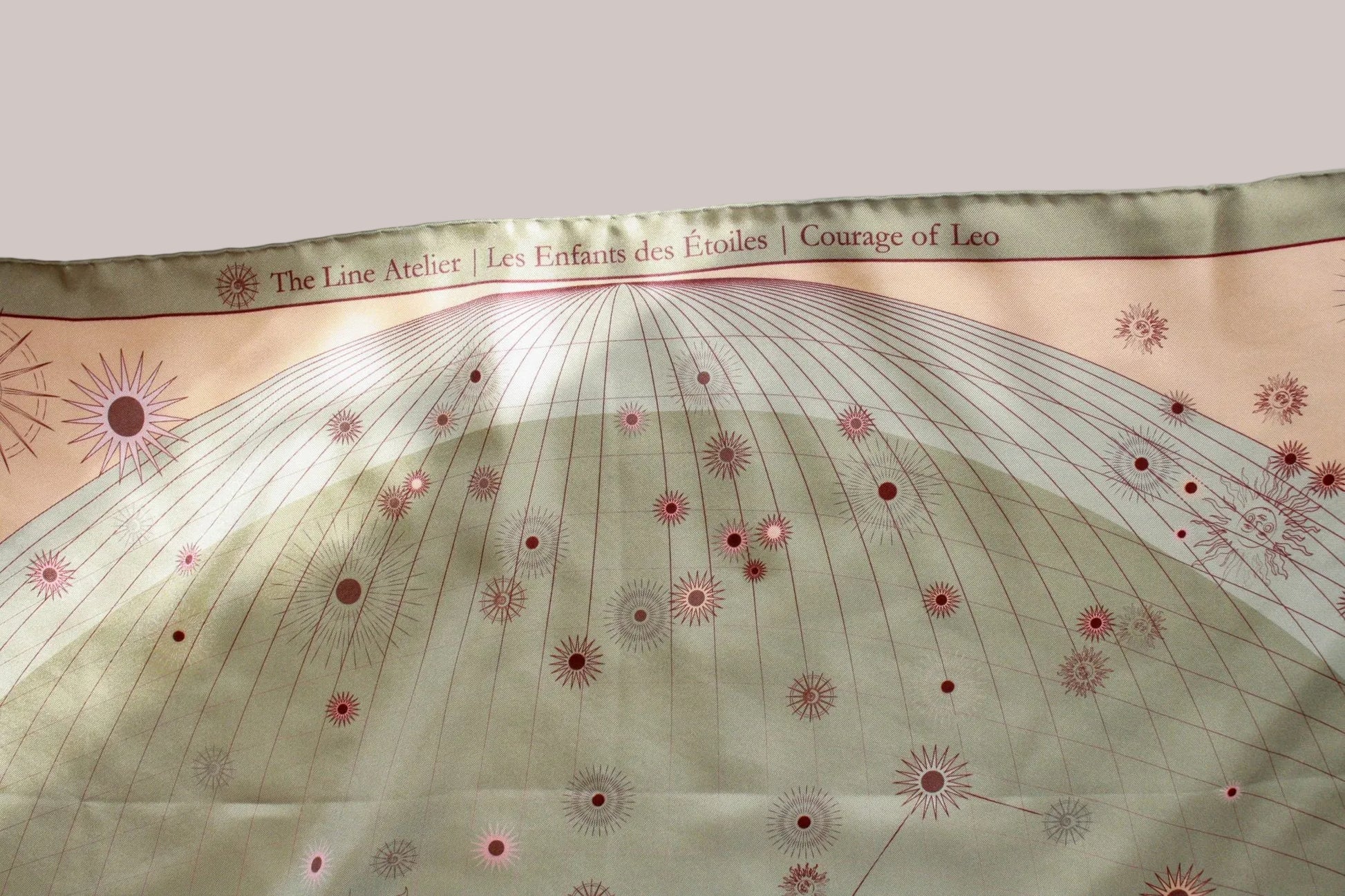 A close-up of a printed scarf or fabric panel with a pale green and beige design, featuring fine radial lines, small pink starburst motifs, and a printed header reading 'The Line Atelier | Les Enfants des Etoiles | Courage of Leo'.