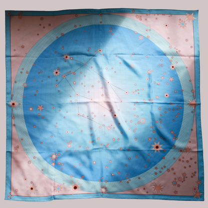 A square silk scarf featuring a celestial design of the Gemini constellation with a large light blue circle, constellation lines, and small stars on a soft pink background, finished with a light blue border and line art of ballet dancers in pairs.