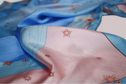 A close-up of a draped silk scarf with a celestial Gemini constellation design of light blue and soft pink, decorated with line art starburst motifs.