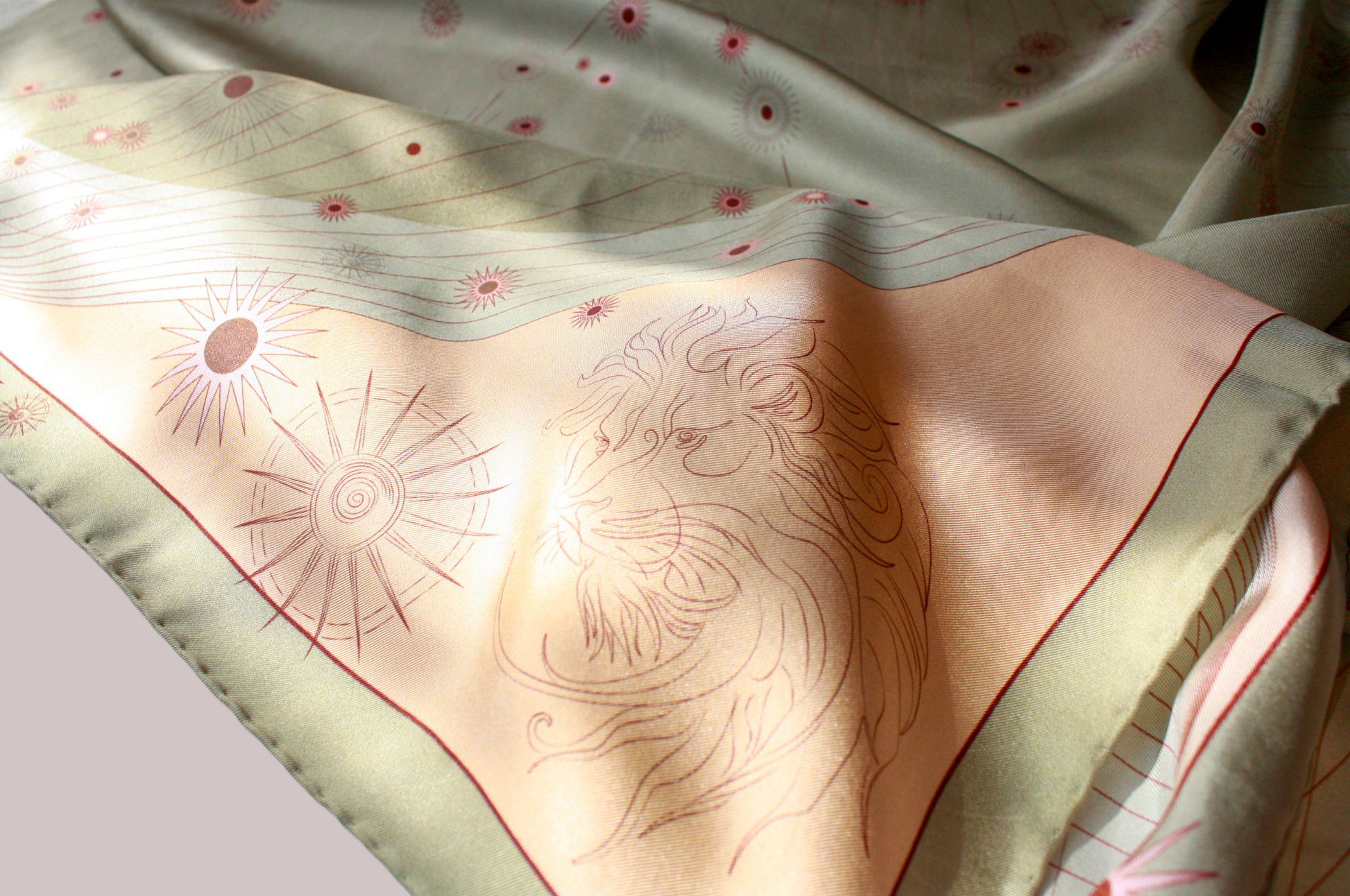 A close-up of a draped scarf featuring a light peach center with a line-art lion and sun motif, bordered by a pale sage green edge with small pink floral accents.