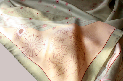 A close-up of a draped scarf featuring a light peach center with a line-art lion and sun motif, bordered by a pale sage green edge with small pink floral accents.