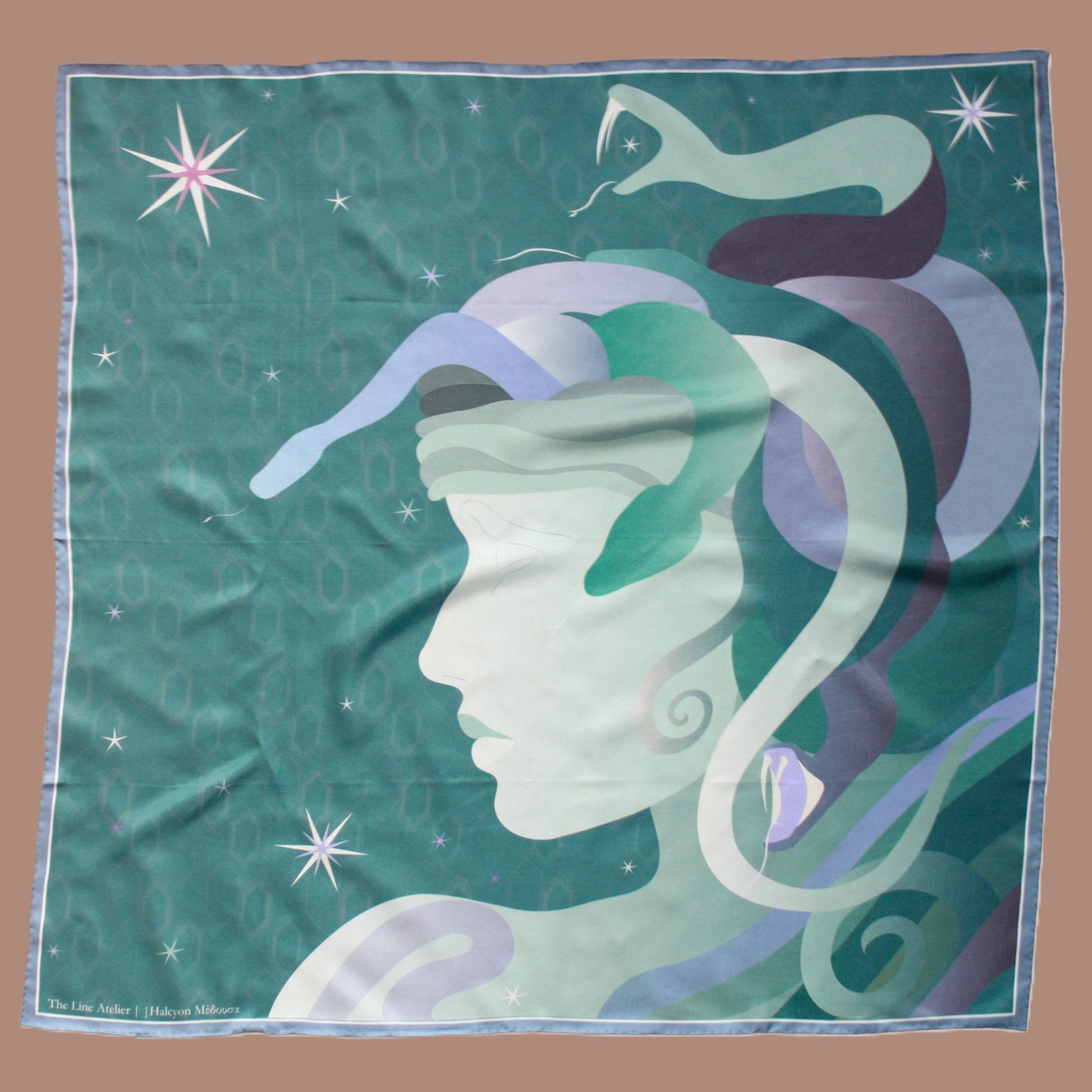 A square silk scarf featuring a stylized profile of Medusa's face in shades of mint green, lavender, and purple, with snakes and stars on a teal background. The scarf is displayed flat against a solid tan background.