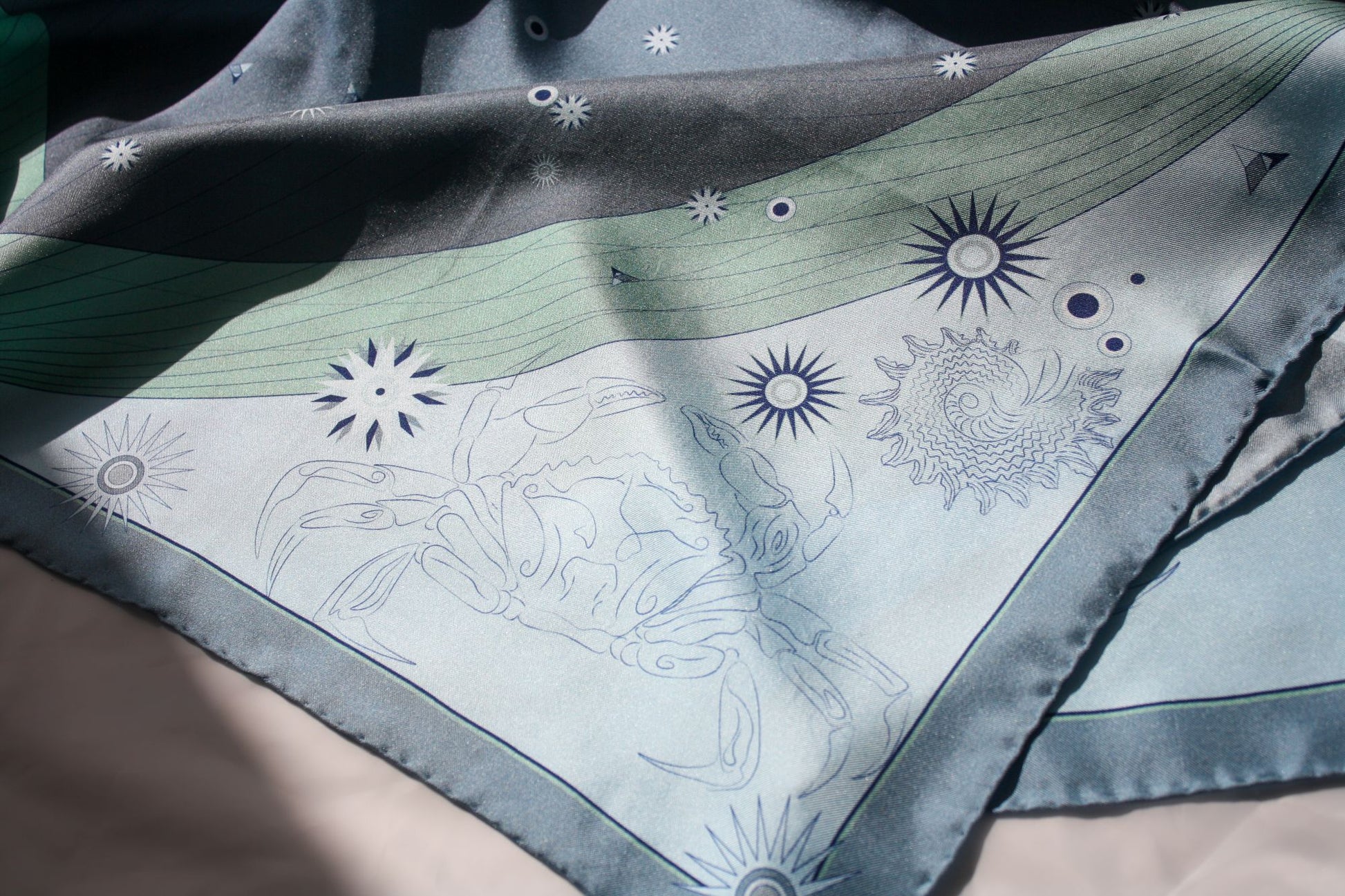 A close-up of a square scarf with a celestial print, featuring a light blue and green background with suns, moons, and a crab illustration.