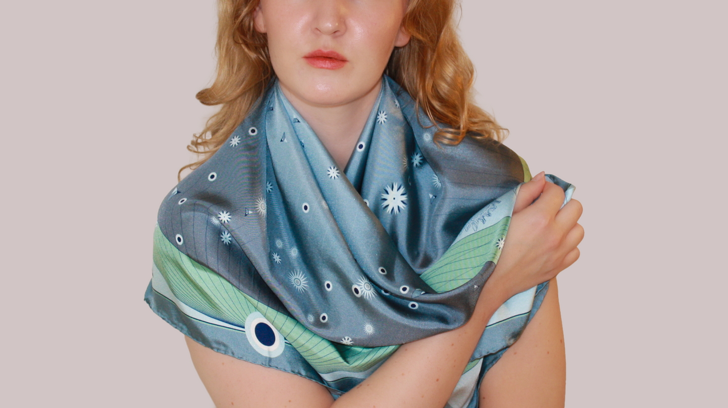 A woman wearing a blue-green Gentle Caner constellation silk scarf with white starburst patterns and green striped borders, shown against a solid beige background.