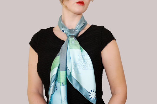 A woman wearing a blue-green Gentle Caner constellation silk scarf with white starburst patterns and green striped borders, shown against a solid taupe background.