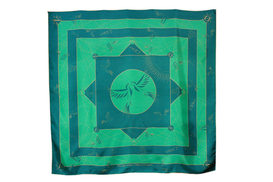A large square silk scarf, Halcyon Compass by The Line Atelier, with a green and teal geometric pattern featuring a central bird motif, hanging on a brown wall.