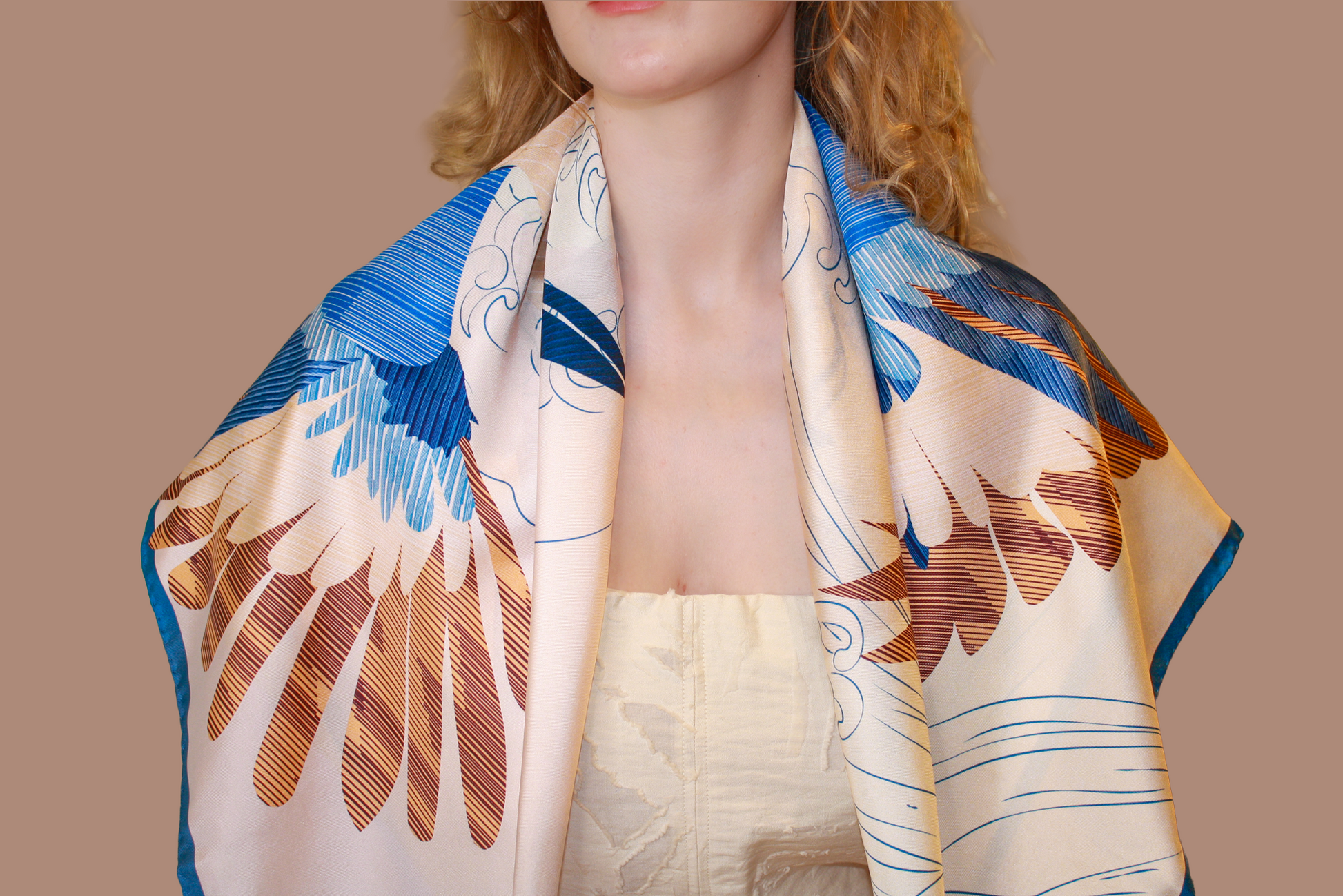 A close-up of a model wearing a cream-colored silk scarf Halcyon Seas with a blue and brown feather pattern, styled over her shoulders.