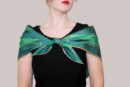 A woman wearing a green silk scarf, Halcyon Compass by The Line Atelier, with gold celestial patterns and text, tied around her neck over a black top, against a solid tan background.
