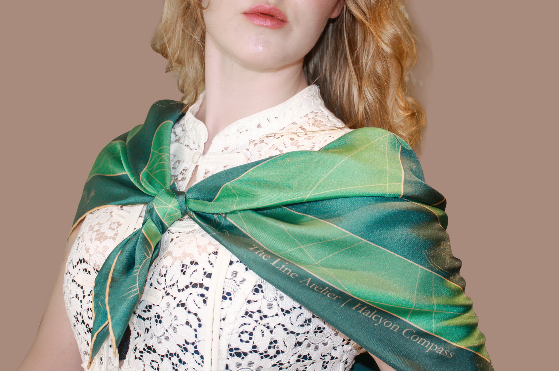 A close-up of a woman wearing a green silk scarf with a map pattern and gold lines tied around her neck over a white lace top, against a solid tan background. The scarf features the text 'The Line Atelier | Halcyon Compass'.