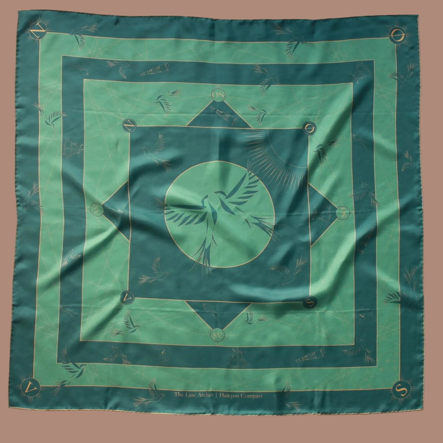 A square silk scarf laid flat on a white background, featuring a green and teal geometric border compass design with a central bird motif inside a circle.