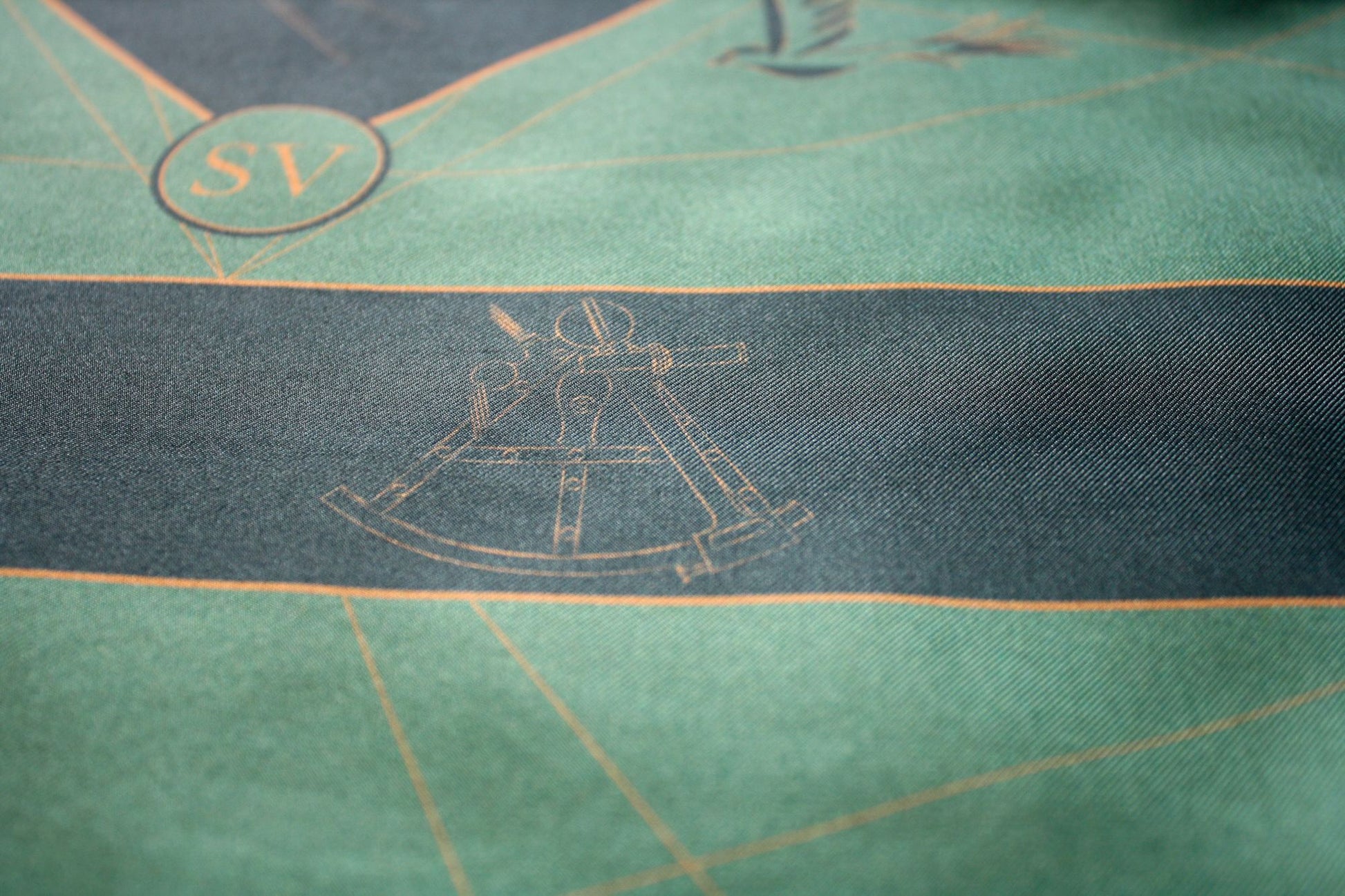 Close-up of a green textile surface with a dark horizontal stripe, fine gold linework, and a faint nautical-style instrument illustration.
