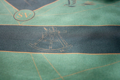 Close-up of a green textile surface with a dark horizontal stripe, fine gold linework, and a faint nautical-style instrument illustration.