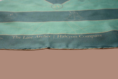 A close-up of a teal and dark teal silk scarf with gold embroidery, featuring the text 'The Line Atelier | Halcyon Compass' and a nautical compass design, laid over a white fabric background.