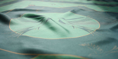 A close-up of a green large silk scarf with a large tonal bird motif outlined in gold and subtle darker green patterning.