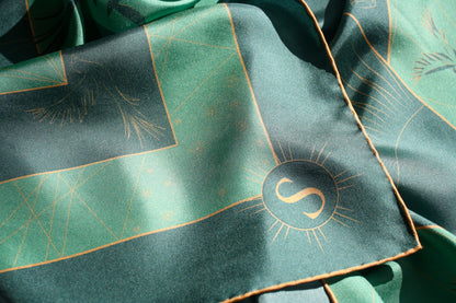 A close-up of a dark green silk scarf with a gold sun and geometric line pattern, featuring a circular compass symbol for South.