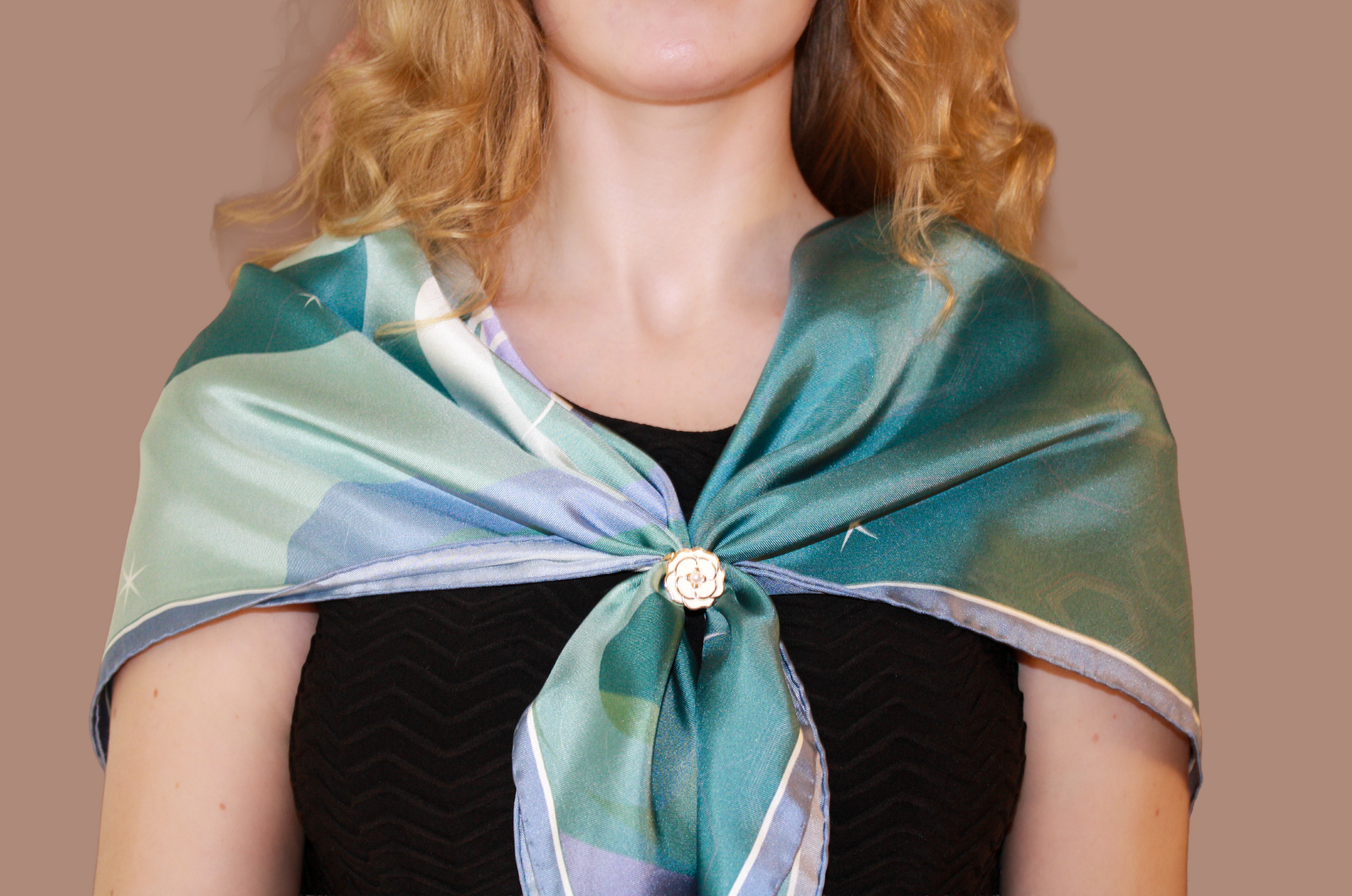 A close-up of a woman wearing a teal and mint green patterned silk scarf, Halcyon Medusa by The Line Atelier, tied around her shoulders, secured with a decorative gold-toned scarf ring, over a black top against a solid tan background.