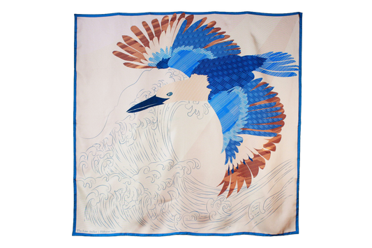 A large square silk scarf, Halcyon Seas by The Line Atelier, featuring a stylized blue and brown bird in flight over white waves, bordered in blue, displayed flat against a brown wall.