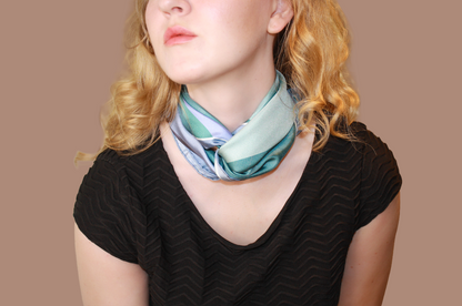 A close-up of a woman wearing a light blue and mint green striped silk scarf, Halcyon Medusa by The Line Atelier, tied around her neck, paired with a black chevron-patterned top, set against a solid warm brown background.