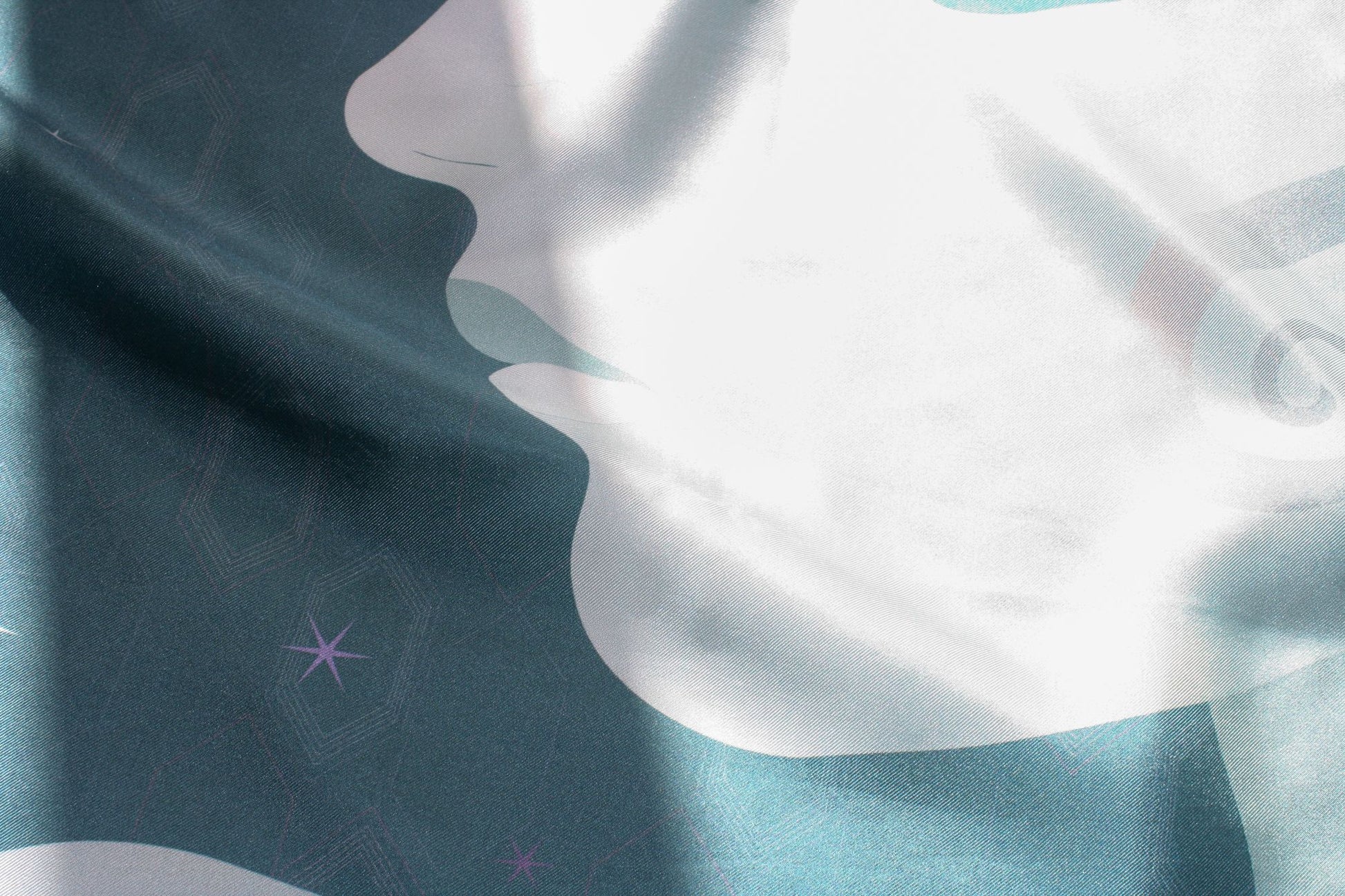 A close-up of a draped silk scarf with a modern abstract design featuring a white silhouette profile of a face against a dark teal and light blue background with subtle geometric patterns and small star details.