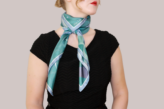 A close-up of a woman wearing a light blue and mint green striped silk scarf, Halcyon Medusa by The Line Atelier, tied around her neck, paired with a black chevron-patterned top, set against a solid warm brown background.