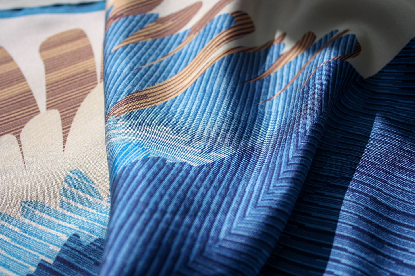 A close-up of a patterned silk scarf with layered blue and navy chevron-like stripes, tan wavy accents, and cream sections, shown in soft folds.