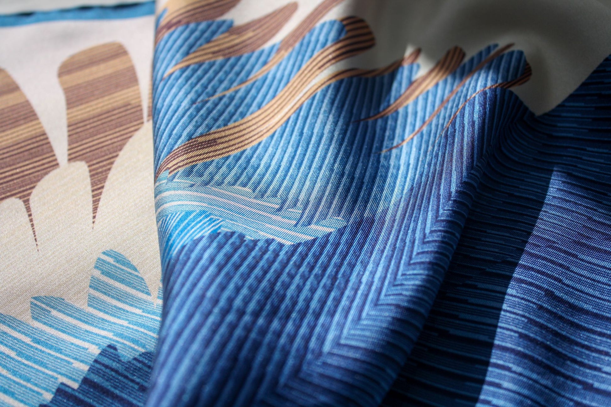 A close-up of a patterned silk scarf with layered blue and navy chevron-like stripes, tan wavy accents, and cream sections, shown in soft folds.
