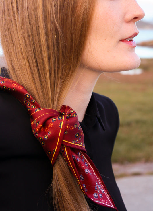 Close-up of a woman's hair with Hearts Ruby silk scarf by The Line Atelier, diamond size, with gem toned pansy flowers on a  blurred Scandinavian coast background
