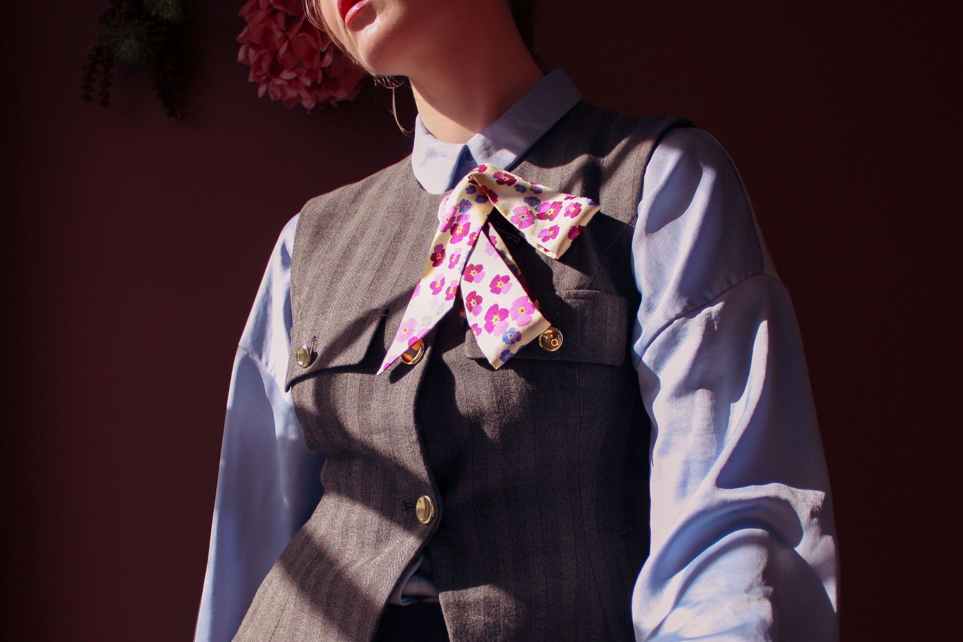 Person wearing a gray vest over a blue shirt with the pink floral scarf Hearts Ease silk twilly by The Line Atelier around the neck against a dark background