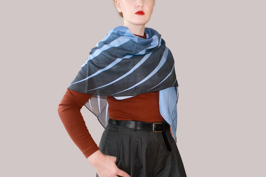 A woman wearing a blue and light blue striped shawl, Ice Valley silk sand wool by The Line Atelier, draped over her shoulders, styled with a rust-brown mock-neck top, a black belt, and dark gray trousers, set against a solid beige background.