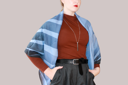 A woman wearing a blue and light blue striped shawl, Ice Valley silk sand wool by The Line Atelier, draped over her shoulders, styled with a rust-brown mock-neck top, a black belt, and dark gray trousers, set against a solid beige background.