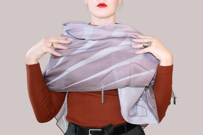 A woman wearing a blue and light blue striped shawl, Love Potion nr 9 silk sand wool by The Line Atelier, draped over her shoulders, styled with a rust-brown mock-neck top, a black belt, and dark gray trousers, set against a solid beige background.