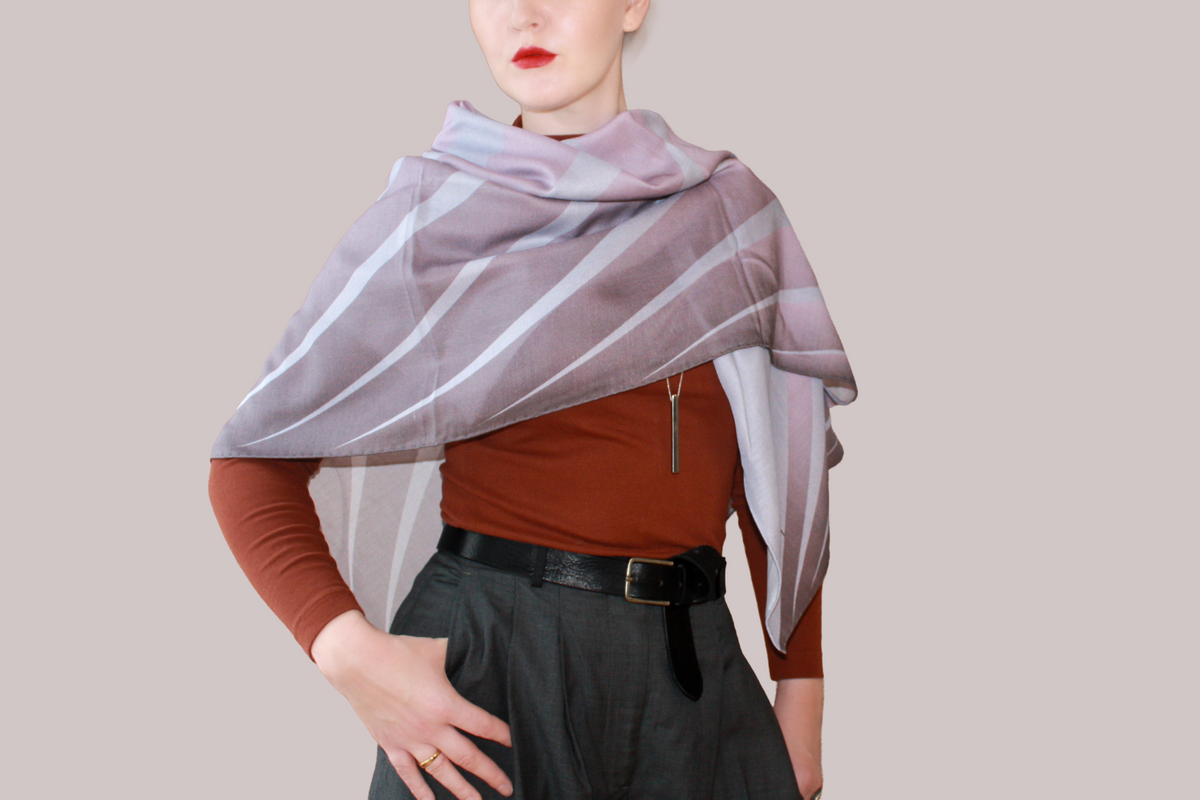 A woman wearing a blue and light blue striped shawl, Love Potion nr 9 silk sand wool by The Line Atelier, draped over her shoulders, styled with a rust-brown mock-neck top, a black belt, and dark gray trousers, set against a solid beige background.