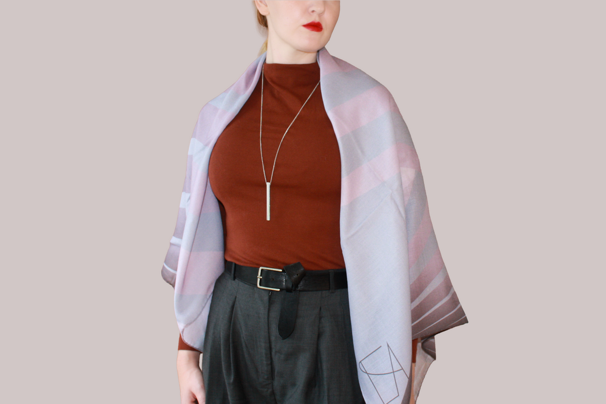 A woman wearing a blue and light blue striped shawl, Love Potion nr 9 silk sand wool by The Line Atelier, draped over her shoulders, styled with a rust-brown mock-neck top, a black belt, and dark gray trousers, set against a solid beige background.