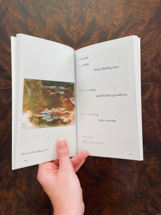 Open Of Shallows & Depths Poem Journal with a Monet painting on one page and text on the other, held by a hand against a wooden background.