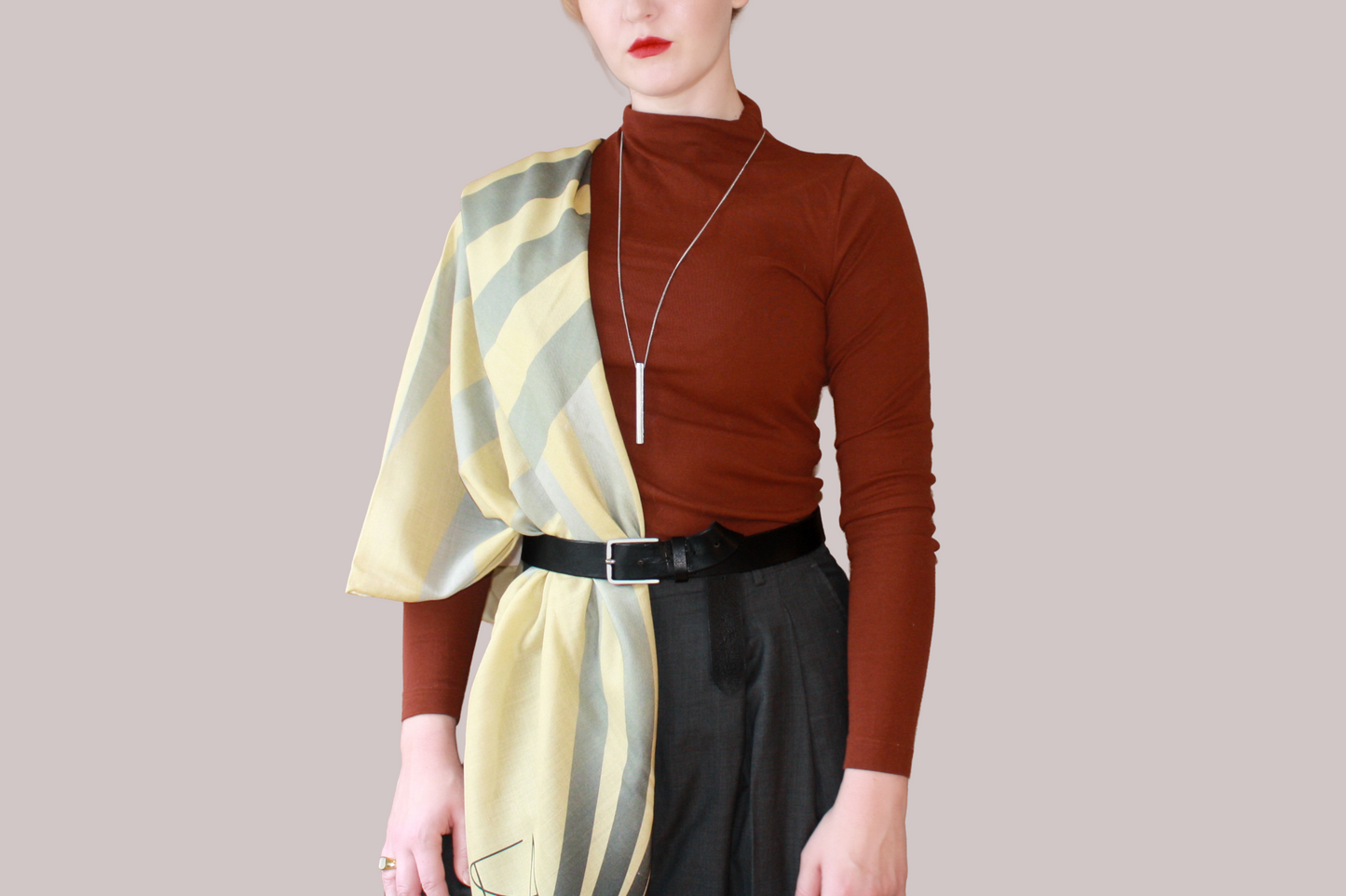 A woman wearing a blue and light blue striped shawl, Willow Grove silk sand wool by The Line Atelier, draped over her shoulders, styled with a rust-brown mock-neck top, a black belt, and dark gray trousers, set against a solid beige background.