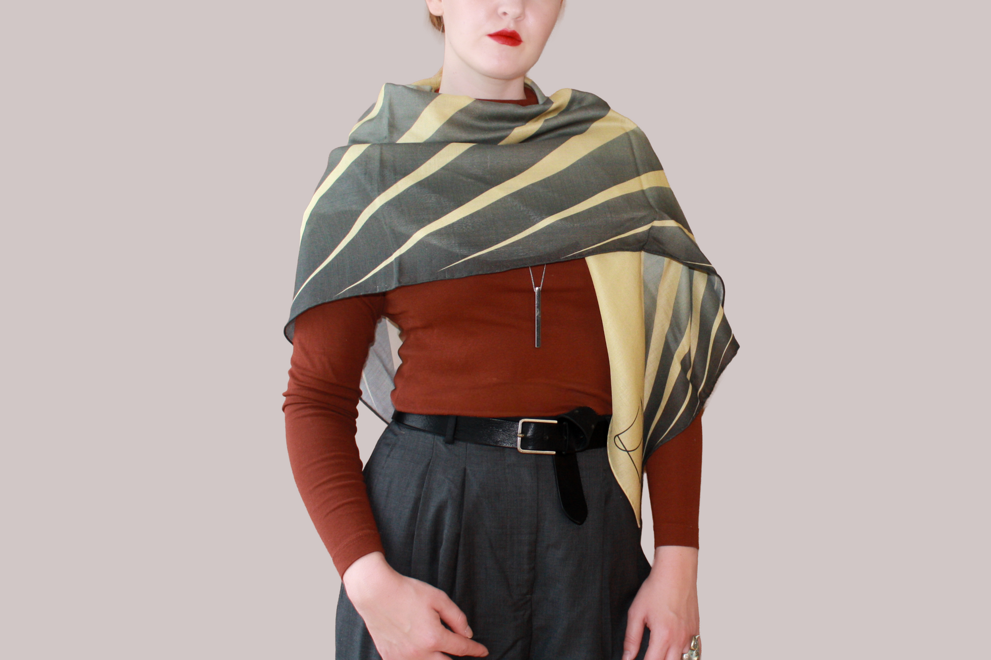 A woman wearing a blue and light blue striped shawl, Willow Grove silk sand wool by The Line Atelier, draped over her shoulders, styled with a rust-brown mock-neck top, a black belt, and dark gray trousers, set against a solid beige background.