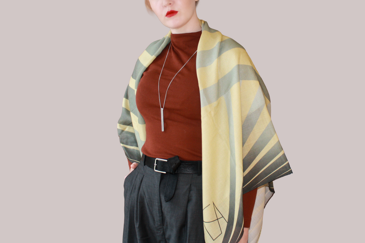A woman wearing a blue and light blue striped shawl, Willow Grove silk sand wool by The Line Atelier, draped over her shoulders, styled with a rust-brown mock-neck top, a black belt, and dark gray trousers, set against a solid beige background.